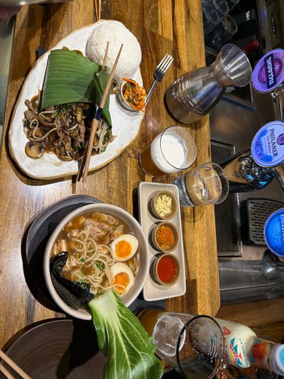 The image provides an overhead view of a wooden dining table within an indoor restaurant or bar establishment located in Pardes Hana - Karkur, Israel. A portion of a person's arm/hand is visible in the upper left corner, suggesting a first-person perspective or the presence of a dining companion.

On the table, a white, irregularly shaped ceramic plate in the upper-mid section holds a dish composed of stir-fried meat (appears to be beef) and mushrooms with green onions, partially encased in a green banana leaf wrapper. A separate mound of white rice and a small white bowl of kimchi are positioned alongside it. Wooden chopsticks rest diagonally across the banana leaf, with a metal fork placed to their right.

Towards the bottom left, a beige bowl contains ramen with noodles, broth, chicken pieces, two halves of a soft-boiled egg with an orange yolk, and a strip of nori seaweed. A head of bok choy is positioned upright next to this bowl. Adjacent to the ramen, a rectangular ceramic tray displays three small white bowls, holding minced garlic, red chili flakes, and a red sauce respectively.

Beverages on the table include a clear glass of light-colored beer with a frothy head, a clear glass of water, and a silver sake carafe. In the background, behind the drinks, multiple beer taps are visible, displaying the brand logos 'Goldstar,' 'Paulaner,' and 'Heineken.' A partially consumed bottle with a colorful label and an empty beer glass are also present in the lower right portion of the image. No other individuals or specific interactions are discernible within the frame.
