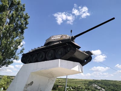 A Soviet-era tank monument is displayed on a concrete pedestal against a bright blue sky with scattered white clouds. The tank, painted olive drab, is positioned with its turret facing left and its long barrel angled upwards. White lettering on the turret reads "ОТ ТРУДЯЩИХСЯ МОЛДАВИИ" (From the workers of Moldova). A red star with a hammer and sickle is visible on the side of the turret. The concrete pedestal is angled and shows signs of wear and tear, with visible cracks and chips. To the left of the tank is the top portion of a tall, leafy tree. In the background, a green, rolling landscape with scattered houses and fields stretches towards the horizon under the clear sky. A person in a green shirt is visible in the lower left corner of the image, near the base of the pedestal.