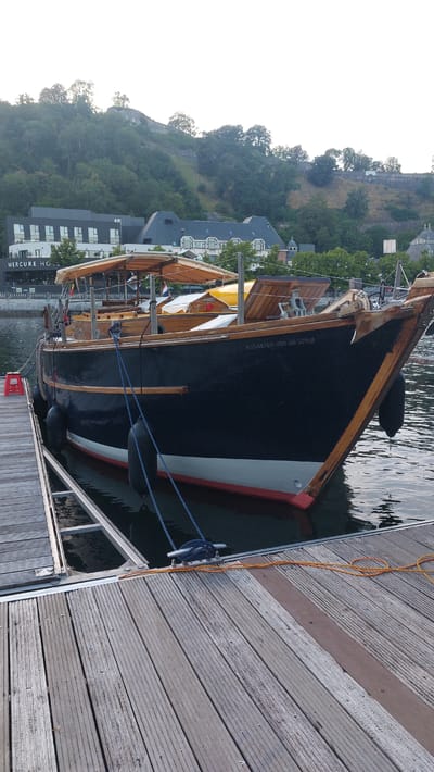 A black, wooden boat named "Kinderen van de Wind" is moored to a wooden dock on a body of water. The boat has a light-colored cabin structure and a wooden deck. A red plastic object is visible on the dock. In the background, a modern building with "MERCURE HO" signage is seen, situated below a tree-covered hill with remnants of a stone wall on its crest. The location is the city of Namur, Belgium, specifically along the river. The scene is captured during the day with overcast or early morning lighting conditions. There are no people visible in the image.