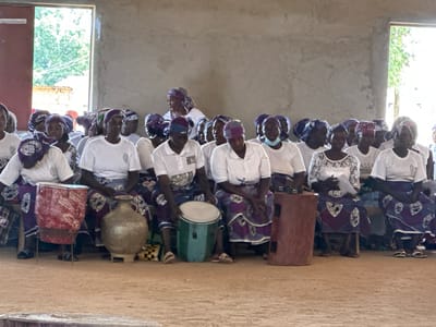A group of women are gathered, seated, in what appears to be an indoor or semi-outdoor setting. The walls are a rough, unpainted concrete, and the floor is packed dirt. Large open doorways reveal greenery and trees outside, suggesting a daytime scene, possibly in a warm climate. The women are dressed in matching white tops and patterned skirts, many with headscarves. They are seated on what seem to be benches or low stools.

In the foreground, several objects are visible that hint at a cultural event or gathering. There is a large, red, cylindrical drum on the left, with white markings. To its right sits a large, metallic, urn-like object. Further to the right, another drum, this one green and cylindrical, is present. Next to it, a tall, reddish-brown cylindrical object, possibly a stool or a large container, is placed.

The women appear attentive, with some looking forward, others engaging with each other. Their expressions are mostly neutral, but the overall atmosphere seems calm and communal. The presence of drums suggests the possibility of music or a performance. The attire and the gathering suggest a community event, possibly a religious service or a celebration, in Kufana, Nigeria. The light filtering in from the doorways indicates it is daytime. Some of the women are wearing masks, a reminder of recent global health concerns.