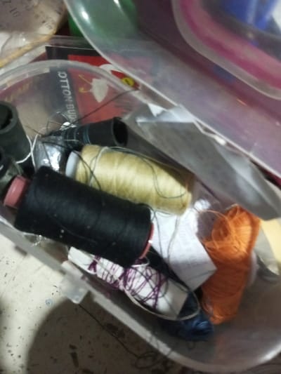 The image displays a close-up, slightly overhead view of various sewing and craft supplies, predominantly spools and bundles of thread, stored in clear plastic containers. The setting appears to be an indoor storage area or workspace, characterized by a somewhat disorganised arrangement of items.

In the center and foreground, several spools of thread are visible. A large black spool with a red core is prominent, lying horizontally. Behind it, a substantial spool of beige or light yellow thread rests. To the right, a vibrant bundle of orange thread is loosely gathered. Towards the bottom left, a white spool or card is visible with purple thread wrapped around it in an irregular pattern. Other smaller black spools and loose white thread strands are scattered throughout the collection.

The threads are housed within, and in some cases spilling out of, clear plastic containers. One such container in the upper left section reveals a label or packaging inside with the text "COTTON BUDS" clearly printed. A small graphic, possibly depicting a cotton swab, is also visible near the text. Other clear plastic containers are stacked or nested above, with faint hints of green and pink visible on their edges.

The lighting is soft, creating mild shadows and reflections on the plastic surfaces and threads. No people are present, and no specific activity is taking place beyond the static arrangement of the craft materials. The background shows a light-colored, indistinct surface, possibly a floor or table.