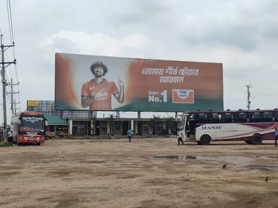 The image shows a bus station in Hatikumrul, Bangladesh. A large billboard advertising "Shah Cement" dominates the background, featuring a man in a sports jersey. In the foreground are two buses, one branded "Sarker Travels" and the other "Hanif." Several individuals are standing around the buses and in the open dirt lot. A one-story building runs along the base of the billboard. Utility poles are visible on the left side of the image. The ground is unpaved and dusty, with puddles of water. The sky is overcast.