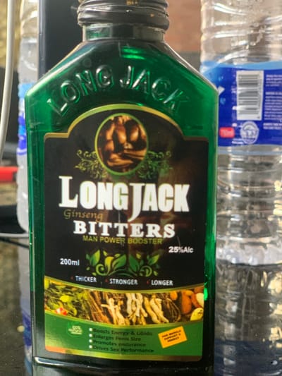 The image displays a close-up, static shot of a dark green, rectangular glass bottle labeled "LONG JACK Ginseng BITTERS MAN POWER BOOSTER" positioned in an indoor environment in Ibeju, Nigeria. The bottle features a prominent black label with white text, indicating "200ml" volume and "25%Alc" alcohol content. The label includes a circular graphic depicting a muscular male torso, accompanied by green leafy motifs. Claims printed on the label include "THICKER", "STRONGER", and "LONGER", along with bullet points stating it "Boosts Energy & Libido," "Enlarges Penis Size," "Promotes endurance," and "Drives Sex Performance." A small yellow sticker on the label reads "FOR MUSCLE SUPPORT & ENERGY," and a green circular icon states "100% Natural." The lower part of the label shows an illustration of various roots and herbs. The bottle cap is not fully visible, but the threaded neck is clear. In the blurred background, several clear plastic water bottles are visible, one to the right bearing a label with a barcode and text, possibly indicating a "750ml" capacity. No individuals are present, and no actions or interactions are depicted. The scene is illuminated by an overhead light source, creating reflections on the bottle's surface.