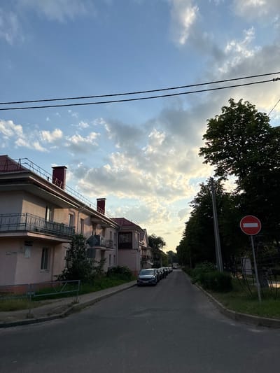 This image depicts a street scene in Zhodzina, Belarus, likely taken in the late afternoon or early evening, judging by the warm, diffused sunlight illuminating the clouds and the shadows. The sky is partly cloudy, with dramatic, illuminated cumulus clouds against a backdrop of blue.

The perspective is from a low angle, looking upwards and along a residential street. On the left side of the frame, the tops of trees and power lines are visible against the sky. To the right, a multi-story building with a pale pink facade dominates the mid-ground. The building has several balconies with decorative metal railings and visible rooflines with chimneys. Further down the street, more buildings and dense green trees line the road.

The street itself is paved and appears to be a quiet residential area. Several cars are parked in a line along the side of the road, suggesting a residential setting. A red and white "no entry" sign is mounted on a pole amidst the trees on the right side of the street, indicating a traffic restriction. There is some grass and vegetation along the sidewalk and around the buildings.

The overall mood is peaceful and serene, capturing a quiet moment in a suburban or town environment. The lighting suggests a pleasant, mild day. There is some faint text visible on a plaque on the building, but it is illegible.
