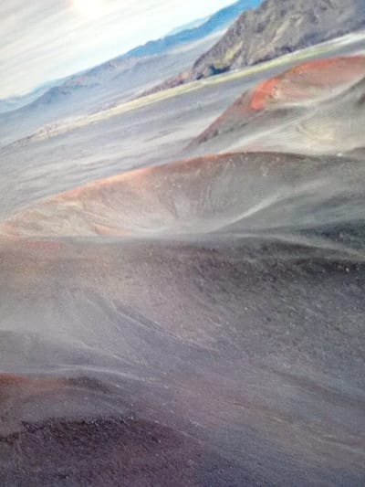 Undulating gray and brown terrain stretches across the frame, punctuated by hints of red and orange hues. Distant mountains fade into the background under a bright sky. The surface appears grainy, possibly depicting a volcanic landscape or arid desert environment. The scene evokes a sense of vastness and desolation.