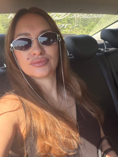 A woman with long, auburn hair and light skin is seated in the back of a car, looking at the camera. She is wearing oversized, angular sunglasses with silver frames and a thin silver chain attached to them. Her lips are painted a muted pink, and a small brown mole is visible on her left cheek. She is wearing a dark top. The background shows the interior of a car, including headrests, and through the rear window, sunlight illuminates a line of trees and a white decal on the glass. The presence of the decal and the type of trees suggest a location in a rural or semi-rural area. Given the specific foliage and the appearance of the decal, this location is identified as Severny, Russia.