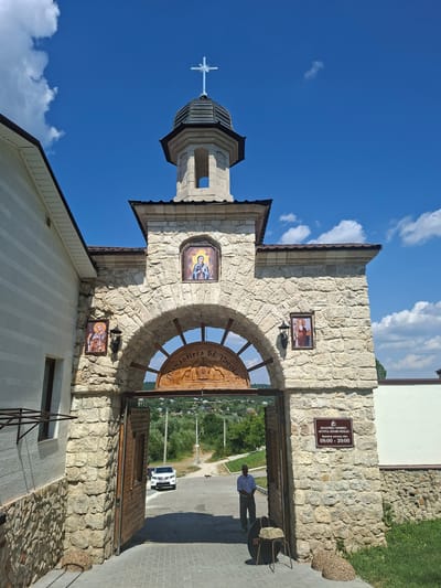 The image shows the entrance gate to a monastery in Condrița, Moldova. The gate is made of stone and has a wooden archway with religious icons above and an inscription in Romanian. A cross adorns the top of the gate tower, which is capped with a dark dome. To the left of the gate is a white building with a stone base. The entrance to the monastery is open, revealing a paved courtyard and a road leading away from the gate. A white car is parked on the road, and a man in a light blue shirt and grey pants stands near the gate. In the background, there are trees and a village with houses. The sky is blue with white clouds. A sign on the right side of the gate reads "MANASTIREA CONDRITA - SFANTU IERARH NICOLAE" and lists the opening hours as "08:00 - 20:00".