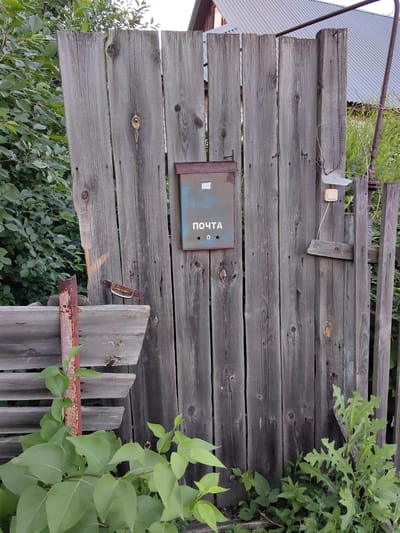 A weathered, gray wooden gate stands partially open, revealing lush green foliage and a glimpse of a corrugated metal roof. Mounted on the gate is a rusty blue mailbox with "ПОЧТА" (POST) printed in white letters. To the right of the mailbox, a beige light switch with a white cover is affixed to the gate. A rusted metal handle for the gate is visible on the left side. The gate is surrounded by dense greenery, with some of the plants encroaching on the structure. The scene appears to be outdoors, likely in a rural or semi-rural setting, potentially in Krestovo-Gorodishche, Russia, given the context provided.