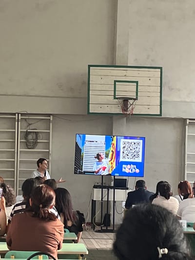 This media file shows a group of people in what appears to be an indoor gymnasium or community hall, possibly in Gladzor, Armenia. The scene is oriented sideways, with a basketball hoop and backboard on the left. To the right of the basketball hoop, there are two screens displaying images and a QR code. The top screen shows a QR code with some blue and yellow graphics below it, and text that reads "FIBA". The bottom screen shows a person in athletic wear, possibly a basketball player, in action.

Several people are visible in the foreground, looking towards the screens. They are seated on benches or in chairs, and their backs are mostly towards the camera. The individuals appear to be engaged with the presentation or broadcast on the screens. The lighting suggests it is daytime, and the setting is indoors with a neutral-colored wall. The overall atmosphere seems to be one of observation and engagement with the presented content.