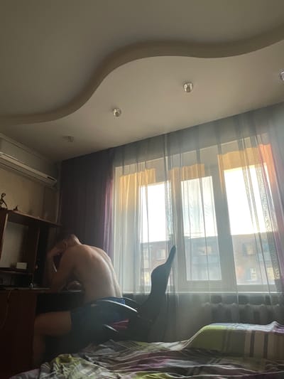 The image depicts an indoor scene, likely a bedroom or study, in Spassk-Dal'nij, Russia. In the foreground, a bed is visible with a striped comforter. A man, shirtless and wearing dark shorts, is seated at a desk on the left side of the frame. He appears to be looking down, with his head resting on his hand, suggesting contemplation or fatigue. To the right of the man, a dark office chair is positioned, with its backrest extending upwards. Behind the desk and man, a window is draped with sheer curtains, allowing soft, diffused sunlight to illuminate the room. The curtains are layered, with sheer white panels over a warmer, orange-hued fabric closer to the window panes. Through the translucent curtains, a brick building with multiple windows is faintly visible, indicating an urban setting. An air conditioning unit is mounted on the wall above the desk. The ceiling features a decorative, wavy design with several recessed lights. The overall lighting suggests it is daytime, possibly late afternoon, given the warm tone of the sunlight.