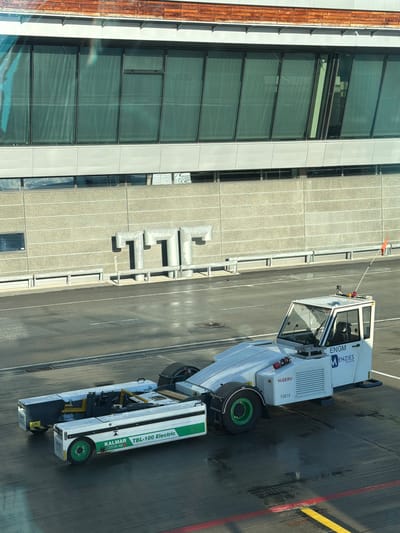 This is a medium shot, taken from a slightly elevated viewpoint, of an airport tarmac at Oslo Gardermoen Airport in Gardermoen, Norway. A white Kalmar TBL-100 Electric tow tractor is parked on the wet asphalt. The tractor is attached to a white and green baggage trailer. The tractor has "ENGM MENZIES AVIATION" printed on its side, along with "HISERV" and "F5814". A small orange flag is attached to a pole on the roof of the tractor. In the background, a modern airport terminal building is visible, with large green-tinted windows and a light grey concrete facade. Several large, tubular metal structures, resembling a series of connected pipes or ducts, are mounted on the concrete wall. The ground is a dark asphalt surface with some visible water puddles and painted yellow and red lines. The lighting suggests it is daytime with a clear sky. There are no people visible in the image.
