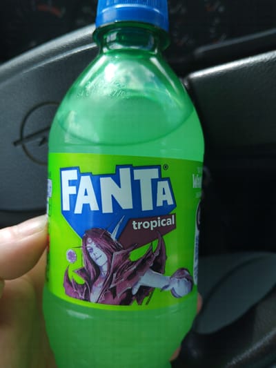 The image shows a bottle of green Fanta Tropical soda with a blue cap. The label features a cartoonish dragon-like character. The bottle is being held by a person's hand and is resting on a car dashboard. The location, Pirdop, Bulgaria, is inferred as a possibility based on the presence of a Bulgarian language version of Fanta Tropical. The date code on the bottle appears to be 21/09/26.