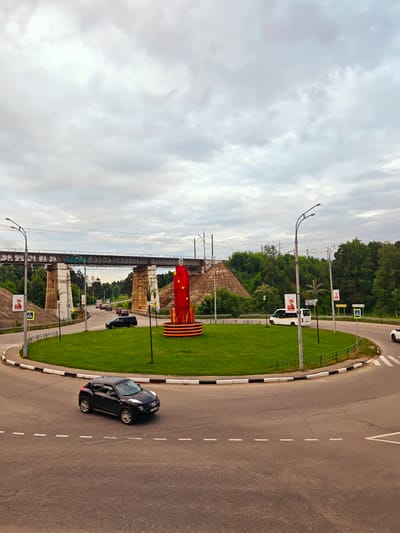 The image depicts a roundabout in Khotkovo, Russia, with a railway overpass in the background. A red, cylindrical monument with gold stars and a St. George's ribbon motif is centrally located on the roundabout's grassy island. Cars are navigating the roundabout, including a black SUV in the foreground and a white van on the road. The sky is overcast with clouds. The environment appears to be a suburban or semi-urban area with trees and vegetation surrounding the roadway.