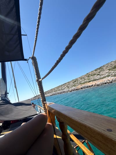 This image depicts a serene scene from a boat sailing on the turquoise waters near Kounoupi, Greece. The perspective is from someone seated on the boat, with their legs visible in the foreground. A thick, braided rope runs diagonally across the frame, attached to the boat's rigging. To the left, a dark fabric canopy covers part of the boat, and a mast is visible. Further back, a person stands near the railing, looking out at the water. The right side of the image showcases a rocky, sparsely vegetated hillside sloping down to the sea. The water is clear and vibrant blue, indicating a sunny day. The overall impression is one of leisure and relaxation on a scenic voyage.