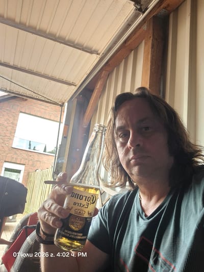 A man with shoulder-length brown hair, wearing a dark green t-shirt, is seated outdoors, holding a cigarette in his right hand and a bottle of Corona Extra beer in his left. The man's face is partially visible, with a neutral expression. The background includes a brick building with a large white-framed window, a wooden fence, and what appears to be a garage or covered patio area with a corrugated metal roof and wooden support beams. A timestamp "01 Iюли 2026 г. 4:02 ч. PM" is overlaid on the lower-left portion of the image. The scene suggests a casual, relaxed setting, possibly a backyard or driveway area in Papenburg, Germany.