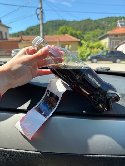 A hand holds a plastic bottle of dark soda, tilted to the right. A sticker with the image of a soccer player named Eduardo Camavinga is attached to the bottle. The bottle rests on a dark surface, likely the dashboard of a vehicle, in front of a blurred background of houses and trees under a bright, sunny sky. The scene appears to be in Beli Osam, Bulgaria, indicated by the context of the image.