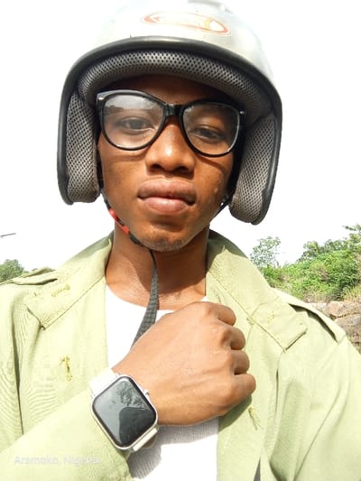 The image shows a young man wearing a silver motorcycle helmet, black framed eyeglasses, a white undershirt, and a khaki green button-up shirt. He is holding his right hand up to his chest, with the back of his hand facing the camera. A silver smartwatch is visible on his left wrist. He has dark skin, short black hair, and a serious expression. His eyes are looking directly at the camera. The background consists of foliage and sky. The image appears to have been taken outside, likely in a natural environment. The image was taken in Iyin, Nigeria, at the coordinates 7.66 latitude, 5.167 longitude. The text "Aramco, Nigeria" appears on the lower left side of the image. The image has a forensic, matter-of-fact description.