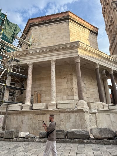 A person stands on a stone sidewalk, looking at their phone. The person is wearing a grey t-shirt and white pants and has a black crossbody bag. Behind the person, a large stone building with columns and architectural details is visible. Part of the building appears to be under scaffolding and covered with a green tarp, suggesting ongoing construction or restoration work. The sky above is partly cloudy, and the lighting suggests it is daytime, possibly late morning or early afternoon. The scene appears to be outdoors in a historic urban setting, given the ancient architecture and stone pavement. The person seems focused on their phone, possibly taking a picture or checking information. The overall impression is of a tourist or visitor exploring a historical site.