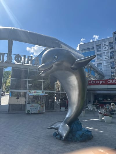 This image depicts a large, metallic dolphin sculpture situated in an urban outdoor setting. The dolphin is positioned in the foreground, facing towards the right of the frame, with its body arcing upwards and its tail resting on a blue, rocky base. The sculpture is highly polished, reflecting the sky and surrounding buildings.

In the background, a modern building with the word "HOTEL" prominently displayed in large white letters is visible. Adjacent to the hotel, a sign with red lettering reads "Burger club". The building features large glass windows, offering a glimpse of the interior and reflecting the outdoor environment. Several tables and chairs are set up outside the building, suggesting an outdoor seating area for a cafe or restaurant. A few people are seated at these tables.

To the left of the dolphin, a tall, metallic archway or structure curves upwards, partially obscuring the sky. The sky itself is a clear blue with a few scattered white clouds. Sunlight is casting shadows on the paved ground, indicating it is daytime. A small kiosk or cart with the text "mininmelts" and "МОРОЗИВО В ГРАНУЛАХ" (which translates to "ice cream in granules") is visible near the dolphin sculpture.

The overall scene suggests a public space, possibly a promenade or a plaza outside a hotel and commercial establishment. The dolphin sculpture serves as a prominent decorative element. The weather appears pleasant, with bright sunlight. The presence of outdoor seating and a kiosk suggests a lively, albeit not crowded, atmosphere.