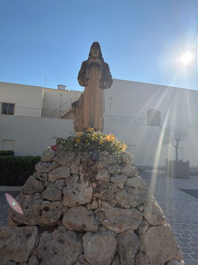 The image depicts a statue of Jesus Christ, hands outstretched and open, possibly in an act of blessing or offering. The statue is made of stone and stands atop a mound of rough, grey rocks. A cluster of yellow and green flowers adorns the rocks near the base of the statue, and a red candle and a dark lantern are visible among the rocks. In the background, a pale cream-colored building with a dark railing and a staircase is present. To the right of the building, a bare tree in a planter is visible. The ground is paved with grey bricks. The sun shines brightly from the upper right corner, casting lens flares across the image and illuminating the scene. The statue and its surroundings appear to be in an outdoor courtyard or plaza. This scene is located in Xagħra, Malta, and is likely a religious monument or shrine.