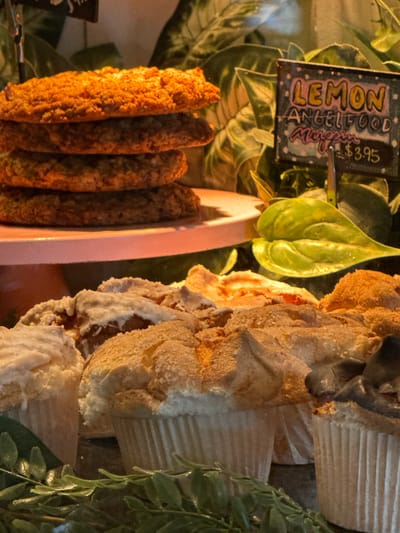 This close-up image, captured in an indoor setting in Oak Brook, United States of America, depicts a vibrant food display, likely within a cafe, bakery, or market. The scene is dominated by various food items arranged amidst lush green foliage, creating an inviting presentation.

On the right side of the frame, several large, individual portions of light-colored baked goods are stacked vertically. These appear to be Lemon Angel Food muffins or cupcakes, characterized by their airy texture, pale, golden-brown crust, and white paper liners. Some portions show a dusting of sugar or fine crumbs on their surface.

To the left, a row of long, cylindrical, golden-brown items is visible. These have a rough, textured exterior, suggesting a savory pastry, fried item, or perhaps a type of breadstick or sausage roll. They are positioned alongside a light pink, curved plastic element, which might be part of a serving tray or display stand.

A small chalkboard sign is prominently displayed in the upper left, providing information about one of the items. The sign features colorful, handwritten text. Clearly legible are "LEMON" in yellow, "ANGEL FOOD" in white, and the partial word "Muffin" in pink script below it. The price "$3.95" is also visible in yellow text.

The background and surrounding areas are filled with green plant life, some appearing to be real broad-leafed plants, and others resembling artificial decorative sprigs, adding a fresh and natural aesthetic to the display. The warm, artificial lighting illuminates the food items, enhancing their textures and colors. No people are visible, and there are no discernible emotions or specific time of day cues beyond the indoor lighting. The activity taking place is the presentation of food for sale or serving.