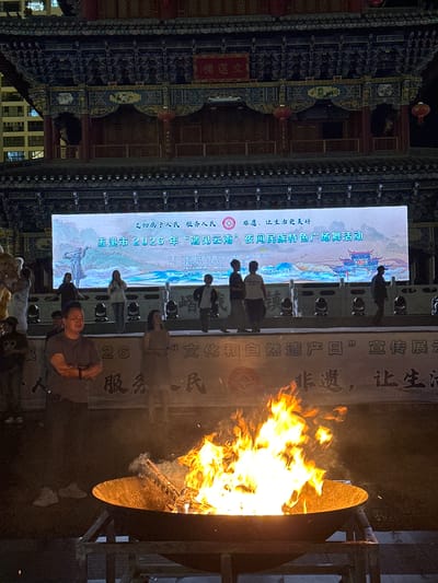 An outdoor nighttime scene features a large bonfire in the foreground, its flames illuminating the immediate surroundings. Behind the fire, a raised platform with stage lighting is visible, with several people standing or walking on it. A large LED screen is mounted on a traditional Chinese-style building, displaying text and an image of a historical Chinese scene with buildings and figures. The building itself is ornately decorated with carvings and red lanterns. To the left of the building, a modern high-rise structure is partially visible. In the foreground, a man in a dark t-shirt is standing with his arms crossed, looking towards the fire. Banners with Chinese characters are displayed in the background, partially obscured by the fire and smoke. The event appears to be taking place in Eshan Yi Autonomous County, China, likely a cultural event given the signage and traditional architecture.