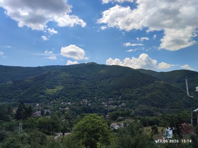 This media file depicts a scenic landscape under a partly cloudy sky. The primary subject is a lush, green mountain range covered in dense forest, with a small village nestled in the valley below. The village consists of scattered houses with red and white roofs, some interspersed with larger, more modern buildings. Trees and vegetation are abundant throughout the scene, with a prominent large tree in the foreground. A power line pole is visible on the left side of the image. In the distance, part of a long, elevated structure, possibly a viaduct or conveyor belt, snakes across the mountainside. The overall setting is rural and mountainous.

The image appears to capture a peaceful, daytime scene. The sun is casting shadows, suggesting it is either late morning or early afternoon. The clouds indicate fair weather.

Visible text in the lower right corner reads "19.06.2026 15:14", indicating the date and time the image was captured. While no people are explicitly visible, the presence of the village implies human habitation and activity. The overall mood conveyed is one of natural beauty and tranquility. The location context provided is Tompsan, Bulgaria.
