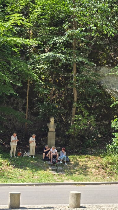 The image depicts a group of people resting outdoors in a lush, green environment, likely a park or natural area, near a road. A stone bust or statue is visible, suggesting a historical or memorial site. The people are dressed casually, indicating a relaxed atmosphere. Some are sitting on a stone ledge, while others are standing. One person is holding a camera, possibly taking photos. The scene appears to be taking place during the daytime, with bright sunlight casting shadows. The presence of trees, foliage, and a rocky incline suggests a natural setting. The location is identified as Vavřinec, Czechia.