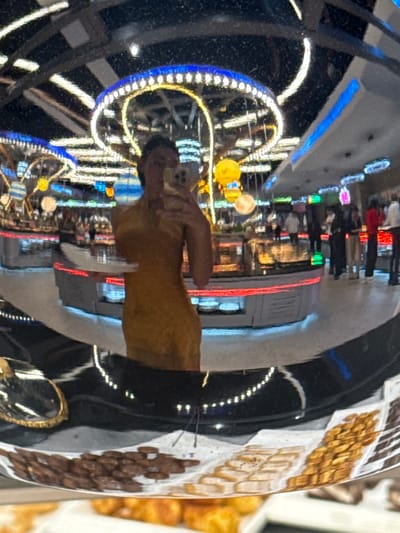 The image shows a distorted reflection in a black, possibly metallic surface. The reflection reveals a person in a yellow shirt taking a picture with their phone. Behind the person, illuminated lights decorate the surrounding area. Various food items appear on the right side.