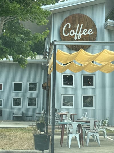 The image shows the exterior of a coffee shop named "Coffee" in Magnolia, United States of America. The building is constructed with corrugated metal siding and features multiple windows. A circular wooden sign with "Coffee" written in white cursive letters is mounted on the upper portion of the building. Yellow fabric shades are draped above the windows, creating an awning-like effect. Outdoor seating with tables and chairs is visible on the patio in front of the coffee shop, some of which are occupied by patrons. Potted plants are placed on the ground near the entrance. The scene is captured during daylight hours.