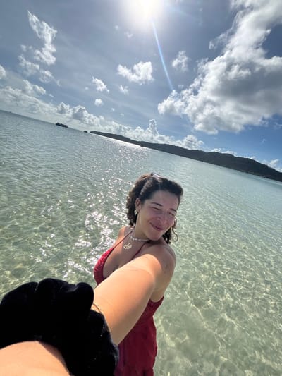 A woman in a red bathing suit stands in shallow, clear ocean water. She is taking a selfie and smiling with her eyes closed. Her arm, holding the camera, is in the foreground, with a black scrunchie on her wrist. The water is calm, with sunlight reflecting off the surface, creating shimmering patterns. In the distance, a lush, green island coastline is visible, with boats sailing on the horizon. The sky is blue with scattered white clouds, and the sun is shining brightly. The location is likely a beach in Preah Sihanouk, Cambodia, known for its beautiful coastlines and islands.