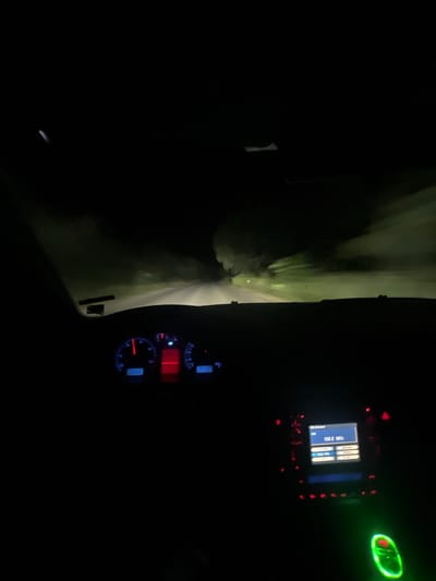 The image depicts the interior of a car driving at night, presumably on a road near Svetlina, Bulgaria. The dashboard lights are illuminated, showing a speedometer with a red needle pointing to approximately 60 km/h, and a radio display showing "108.0 MHz". The exterior view through the windshield is dark, with faint headlights illuminating a misty, tree-lined road ahead. The atmosphere suggests driving in fog or a similar atmospheric condition. There are no people or animals visible in the frame.