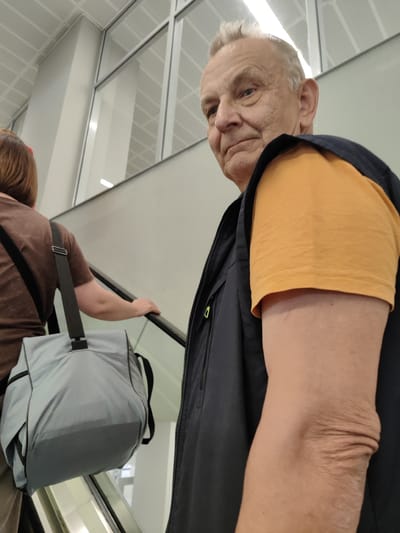 A medium shot captures a man in his late 60s or early 70s with short, graying hair, looking directly at the camera. He is wearing an orange t-shirt and a dark vest over it. His left arm is prominently featured, showing aged skin and wrinkles around the elbow. He appears to be on an escalator. Behind him, a woman with brown hair is also on the escalator, carrying a light gray shoulder bag. The environment appears to be a modern indoor space with large windows and white ceiling tiles, possibly an airport terminal or a shopping mall. The location is identified as Khrabrovo, Russia.