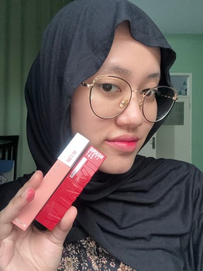 A close-up image, captured indoors in Patrang, Indonesia, features a young adult female from the chest up. She wears a black hijab, a black top with a subtle floral pattern on the upper left, and round, gold-rimmed eyeglasses. Her lips are colored with a reddish-pink cosmetic. Her right hand, with red-painted fingernails, holds two cosmetic tubes towards the camera: one light peach-colored tube labeled "MAYBELLINE MATTE INK" and one red, textured tube labeled "MAYBELLINE SUPER STAY VINYL INK". The subject maintains eye contact with the camera. The background consists of a light green wall, with a dark furniture piece partially visible on the left and a white door with a light blue geometric pattern on the right.