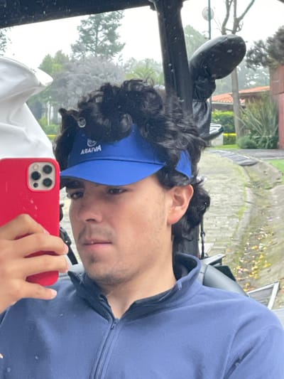 A male individual is depicted taking a selfie with a red smartphone while seated in what appears to be a golf cart within Fraccionamiento y Club de Golf los Encinos, Mexico. He possesses dark, curly hair and light facial stubble, wearing a blue visor with "ARAMVA" printed on it and a dark blue quarter-zip pullover. A black padded golf club head cover is visible on a club mounted to the golf cart's structure behind him. The background reveals an overcast outdoor environment with green foliage, a paved cart path exhibiting scattered leaves, and distant residential-style buildings featuring reddish-brown tiled roofs, consistent with a golf course community. Moisture droplets are present on surfaces, indicating damp conditions.