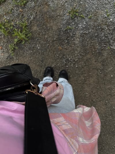 An overhead, first-person perspective photograph depicts the lower body of an individual standing on a gravel path in the city of Susly, Ukraine. The ground consists of gray and brown pebbles mixed with dirt, interspersed with sparse green vegetation, notably on the left side of the frame. The subject wears light blue denim jeans and black, chunky-soled loafers. A plain pink garment is visible beneath a plaid flannel shirt, which features a pattern of pink, white, blue, and yellow-orange lines. The sleeves of the flannel shirt are rolled or pushed up. A black, possibly synthetic leather, bag with a metallic zipper and a gold-colored clasp on its black strap is positioned on the left side of the subject's body. The individual appears to be stationary.