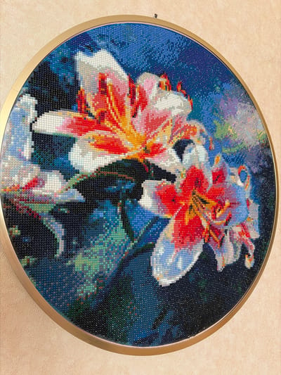 A circular diamond painting depicts a close-up of lilies with vibrant red and white petals against a dark blue background. The painting is framed by a thin, golden metal rim and hangs on a textured, light beige wall. A small black hook is visible above the painting. The scene is indoors, in what appears to be a residential or display setting. No people are present. The location in Мяндиги, Russia is not discernible from the image.