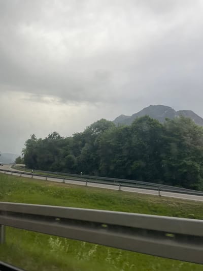 The image captures a view from a vehicle traveling on a highway in Farchant, Germany, on a cloudy day. A guardrail runs along the foreground, with a grassy embankment sloping down to its right. A two-lane road curves to the left, bordered by a second guardrail. Beyond the road, a dense line of lush green trees forms a backdrop, with the imposing, rocky peaks of a mountain range visible in the distance, partially obscured by the overcast sky. The overall impression is one of natural, scenic beauty, typical of the Bavarian Alps region.