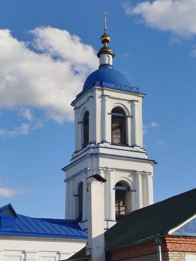 The image depicts a white bell tower with a blue dome and gold cross, standing against a backdrop of a blue sky with white clouds. The architecture features arched openings with columns. The tower rises from a building with a blue roof on one side and a green roof on the other, with portions of a brick wall visible. The location is Мамонтово, Russia. No people are visible.
