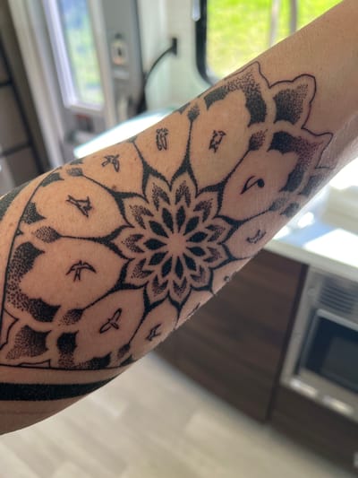 A human forearm displays a large, circular tattoo composed of concentric floral or petal-like designs. The tattoo is predominantly black ink, with outlines and stippling used to create shading and texture. Scattered within the petal shapes are small images of birds in various flight poses, as well as symbols resembling musical notes. The tattoo appears to cover a significant portion of the forearm. The surrounding environment is a brightly lit interior, featuring a window, a partial view of a kitchen cabinet, and laminate flooring. It is not possible to infer the specific location within the city of Enosburgh, United States of America, from the provided image. There are no other persons, actions, or interactions evident in the photo.