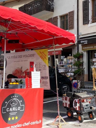 This image depicts a street scene in Abondance, France, featuring a vibrant red market stall under a large red umbrella. The stall, identified by "CHEF CARLO" and "LA POELE DES VOLCANS" banners, displays kitchenware and produce, including zucchini and lemons in a black tray, and stacks of red and white boxes. A person with dark hair is partially visible behind the counter, looking down. A yellow banner with "OFFRE DÉCOUVERTE -20%" is attached to the stall.
To the right of the stall, a black vehicle is parked. Further right, a small, four-wheeled cart, decorated with red and white checkered fabric featuring a white cross, holds sound equipment including a mixer, and a brass trombone is positioned vertically nearby. A "STATIONNEMENT INTERDIT" (No Parking) sign is visible near the cart.
In the background, two multi-story buildings with light-colored facades line the street. They feature windows with closed brown wooden shutters. One building has an ornate dark metal balcony. Ground-level businesses are visible, including a "sherpa ALIMENTATION" store and a "BRASSERIE," with various items displayed outside and potted plants.