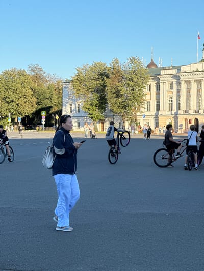 In Yazvitsevo, Russia, on a clear day, a group of young people engage in bicycle stunts in a city square. In the foreground, a woman in a dark blue jacket and light blue pants walks while looking at her phone. She carries a light-colored backpack. Behind her, several teenagers on bicycles are the central focus. One boy performs a wheelie on a black bicycle with purple accents on the wheels. Another boy sits on a red bicycle, facing away from the camera. To the right, a third boy on a black bicycle stands with one foot on the ground. Further in the background, more individuals are seen walking and pushing a stroller. A large, ornate building with classical architecture, possibly a government or cultural institution, dominates the background. Trees line the perimeter of the square, and traffic signs are visible on the left. The ground is paved asphalt. The sky is clear and blue.