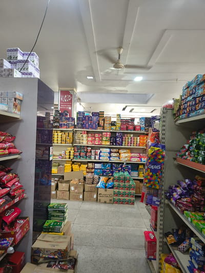 This is a medium shot, interior, eye-level photograph of a grocery store aisle in Sidhpur, India. The aisle is filled with shelves of packaged food items, predominantly snacks and sweets. On the left, shelves are stocked with red and white packaged snacks, and further up, boxes of white and purple packaged beverages. In the center, a tall display of Kit Kat chocolate bars is visible, along with shelves of various biscuits and snacks in yellow and blue packaging. A person's arm is reaching towards the upper shelves in the center. On the right, shelves are filled with a variety of colorful packaged candies and snacks. The floor is made of gray tiles. The ceiling features a white, paneled design with a ceiling fan and recessed lighting. The overall impression is that of a well-stocked convenience store or supermarket.