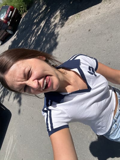 A young woman is photographed from a high angle, outdoors, with a road and shadows visible. She is wearing a white t-shirt with navy blue trim and blue denim shorts. Her eyes are closed, and her tongue is sticking out as she smiles. A maroon car is parked on the left side of the frame, and green foliage is visible in the background. The scene appears to be in the city of Kolašin, Montenegro.