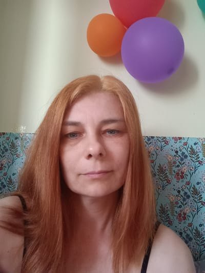 A medium shot photograph captures a fair-skinned woman with long, ginger hair, looking directly at the camera. Her eyes are a light blue, and there are subtle lines around them. Her lips are slightly parted, and her expression is neutral. She is wearing a dark colored strap visible on her left shoulder.
The background features a light green wall. Towards the upper right of the frame, three balloons are visible: an orange balloon on the left, a purple balloon in the center, and a partially visible pink balloon on the far right. Below the balloons, a section of wallpaper with a floral pattern in blue, white, and red is visible. The image appears to be taken indoors, possibly in a residential setting in Vyškov, Czechia.