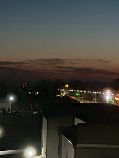 This is a nighttime photograph taken from a high vantage point, likely a building, in Vinnytski Khutory, Ukraine. The scene depicts a twilight sky with gradient hues of blue and orange, transitioning into darker tones as it meets a horizon dotted with distant lights from buildings and trees. In the foreground, the dark silhouettes of rooftops are visible, with some illuminated by artificial lights. A street with a series of bright streetlights stretches across the middle ground, illuminating a road where a few vehicles are present. To the right, a tall, illuminated sign, likely for a business or gas station, stands out against the dark sky. The overall impression is that of a suburban or semi-urban environment at dusk or early night.