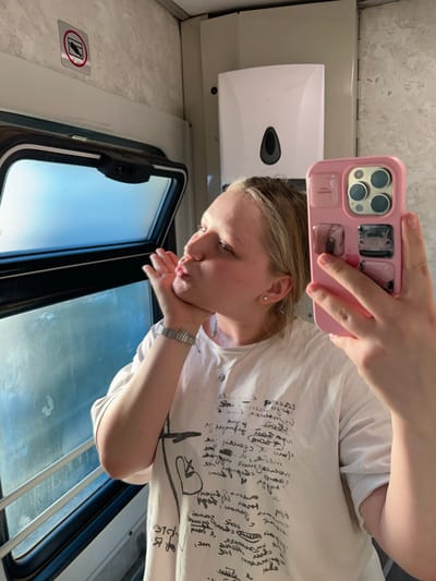 The image captures a selfie taken by a young person inside what appears to be a train or bus, oriented as if viewed from the ceiling looking down, but described here in its intended upright orientation.
The central subject is a light-skinned individual with blonde hair, wearing a white t-shirt adorned with extensive black handwritten-style text, drawings of hearts, arrows, and abstract scribbles. She holds a pink smartphone with a distinct camera array in her right hand, extending it to take the picture. Her left hand is positioned under her chin, and her lips are pursed in a playful "kiss" or "duck face" expression. She wears a silver watch on her left wrist and a delicate silver necklace. A small stud earring is visible in her right ear, and she appears to have braces on her lower teeth.
The background elements suggest a vehicle's interior. To the person's right, a white dispenser with a black oval opening is mounted on a textured, light-colored wall. A window occupies the lower portion of the frame, revealing a bright blue sky and some indistinct reflections, indicating movement and a daytime setting. The window frame is dark with a visible latch mechanism. On the wall beside the window, a silver plaque with a red-circled "no littering" symbol (a hand dropping an item into a bin, crossed out) is clearly visible. The overall mood is casual and lighthearted. The location context of Zimovniki, Russia, further supports a travel setting.