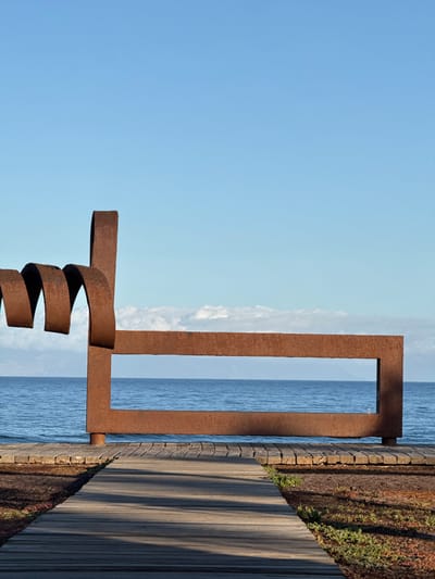 The image depicts a large, rusted metal sculpture situated on a wooden boardwalk overlooking the ocean in Playa De Las Américas, Spain. The sculpture consists of two main elements: a coiled, wavy section on the left and a rectangular frame on the right, both made of weathered, reddish-brown metal. The ocean is a vibrant blue, with gentle waves and a clear horizon. The sky is a pale blue with a few scattered clouds. The boardwalk is constructed of weathered wooden planks, with some areas showing signs of age and wear. To the left of the boardwalk, there is a patch of reddish-brown earth with sparse green vegetation. The scene is devoid of people, creating a sense of peaceful solitude. The lighting suggests it is daytime, with the sun casting shadows across the boardwalk.