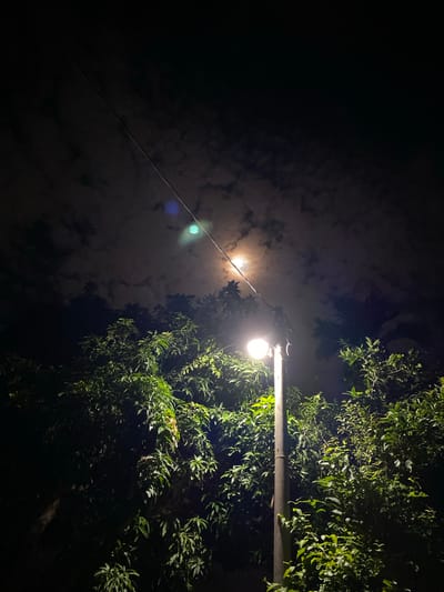 This is a low-angle, outdoor, night shot of a street lamp illuminating a dense cluster of green foliage. The lamp is positioned on the right side of the frame, mounted on a metal pole. A wire extends diagonally from the lamp across the frame towards the left. In the background, a hazy moon is visible through cloudy skies. The overall impression is one of a dimly lit, humid night, possibly in a tropical or subtropical environment. The intense light from the lamp highlights the vibrant green of the leaves, creating a strong contrast with the dark sky and foliage. There are no people or visible text in the image. The location context suggests this scene is in Lambaro Kueh, Indonesia. The time of day is clearly night, and the presence of the moon and clouds indicates it's likely a partly cloudy evening. The lush greenery suggests a natural or semi-natural setting, possibly a garden or park area.