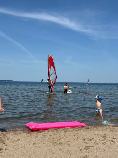 A group of people are windsurfing on a sunny day in Rewa, Poland. The sky is a clear blue with some wispy clouds. The water is calm and shows ripples from the wind. In the foreground, a person is lying on a bright pink inflatable mattress on the sandy beach, with their hand reaching out. Another person is in the water, splashing around. Further out, several people are windsurfing with their sails up. One windsurfer in the middle ground has a red and white sail. The overall mood is relaxed and recreational, suggesting a summer day at the beach.